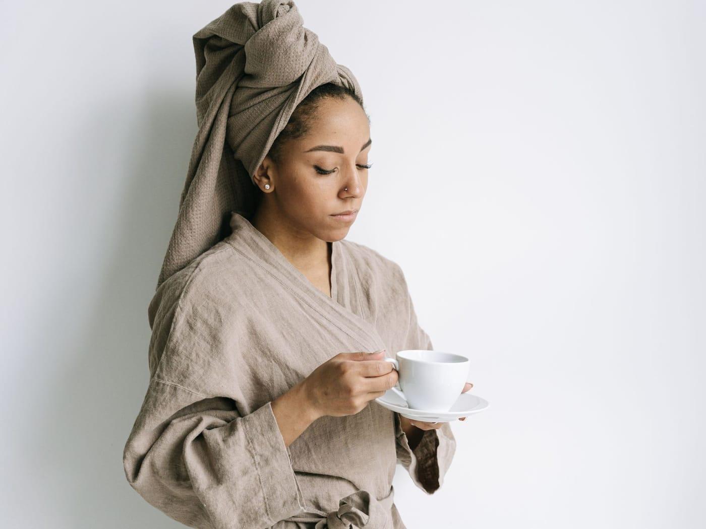 A woman in a linen robe with her hair wrapped in a towel, holding a cup of tea