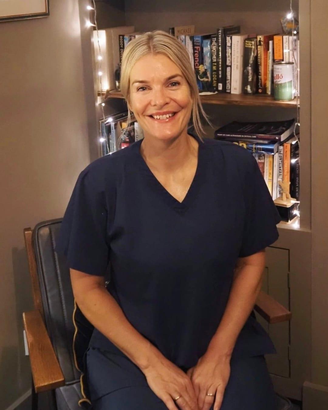 Emma Dutfield, smiling, seated in navy scrubs in front of a softly lit bookshelf