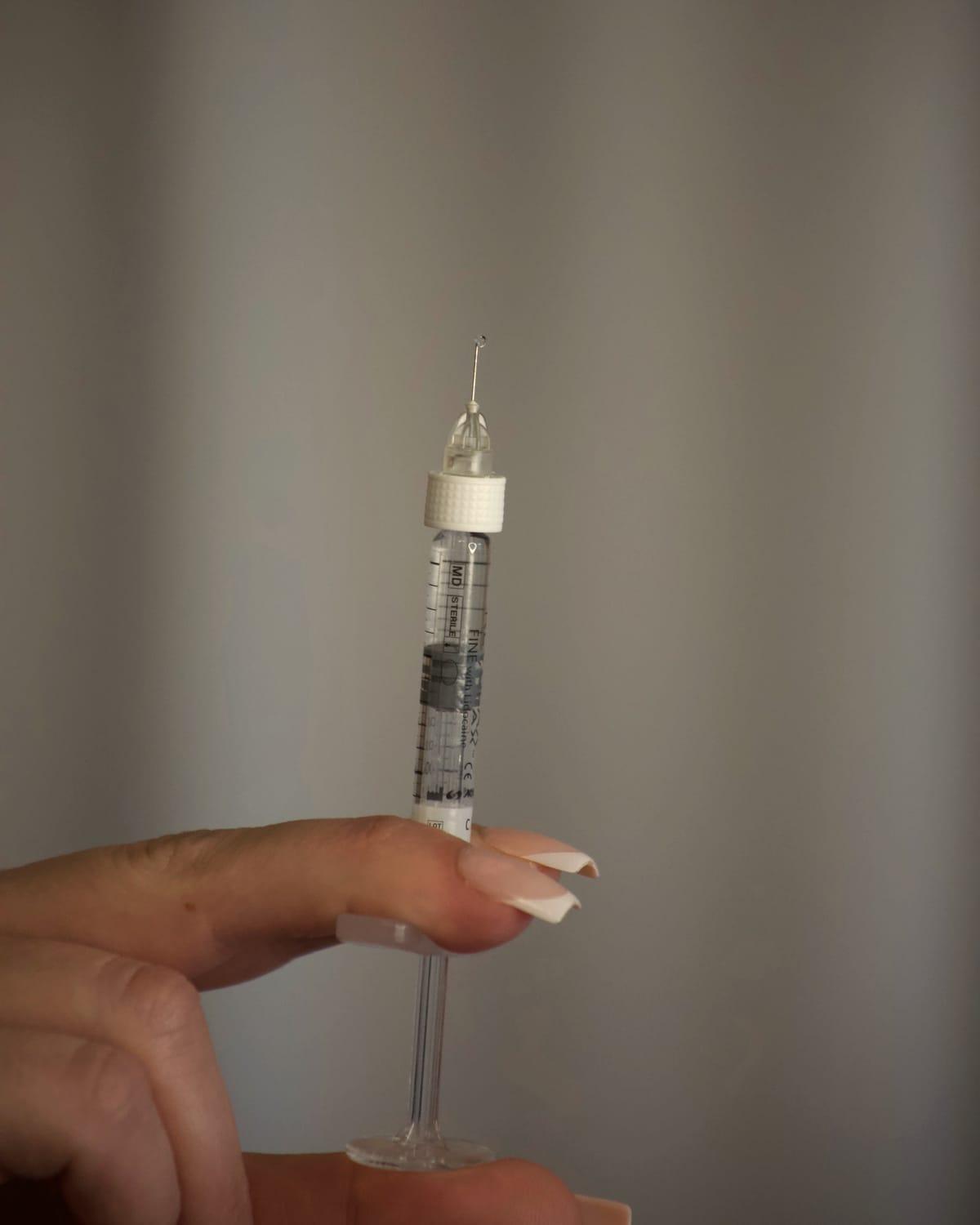 A filler syringe balanced between a practitioner's fingertips against a soft curtain
