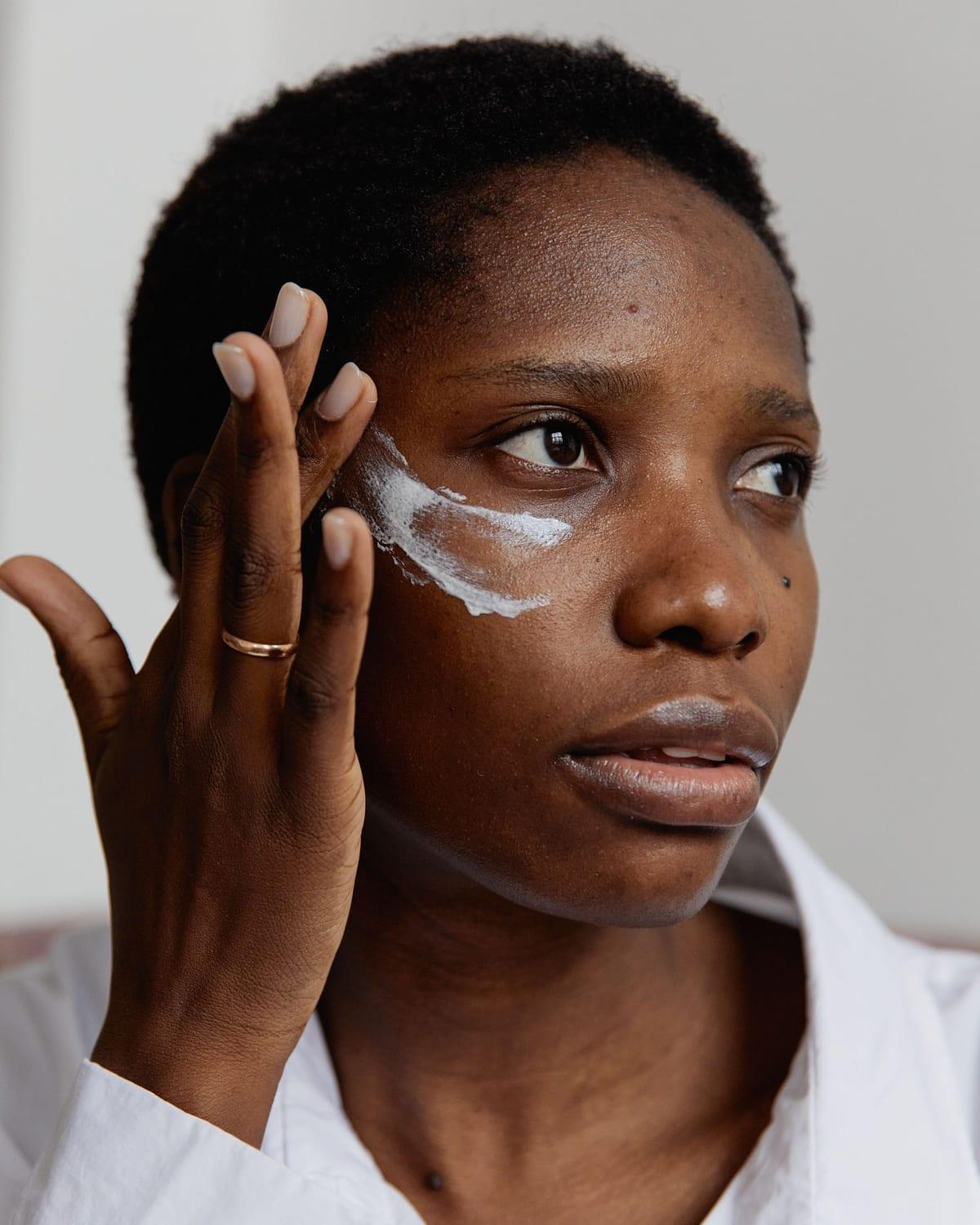 A woman in a white shirt smoothing cream beneath her eye, in soft natural light